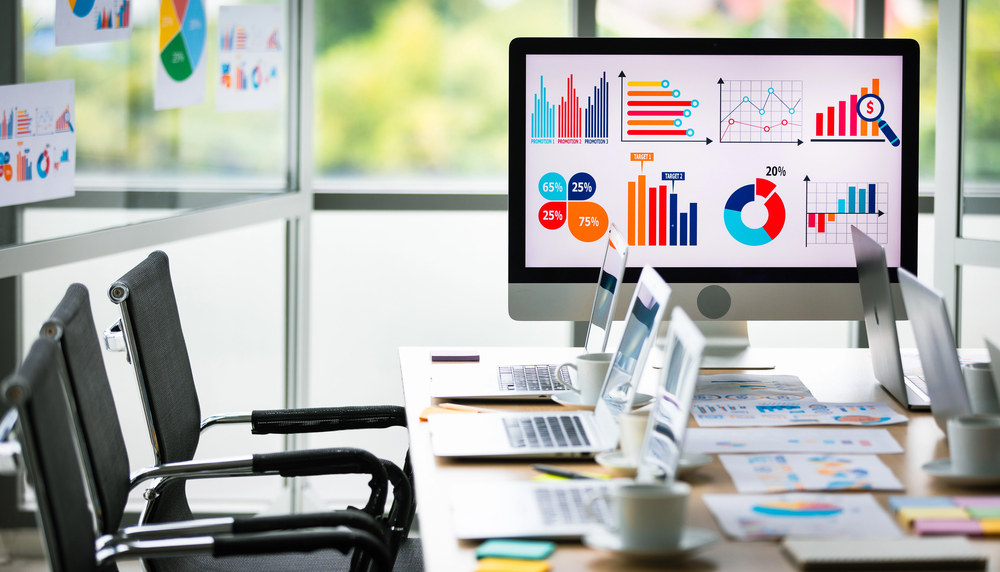 Analysis sales target growth graph chart investment report data on big computer screen monitor placed in middle of meeting table in front glass building windows with garden view in blurred background
