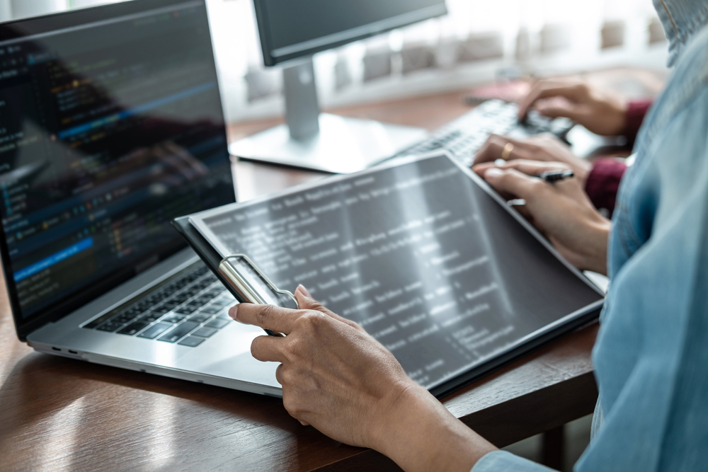 Asian woman programmer holding code data on clipboard and discussion with partner about new project while working together to writing code for new website or application in software development office